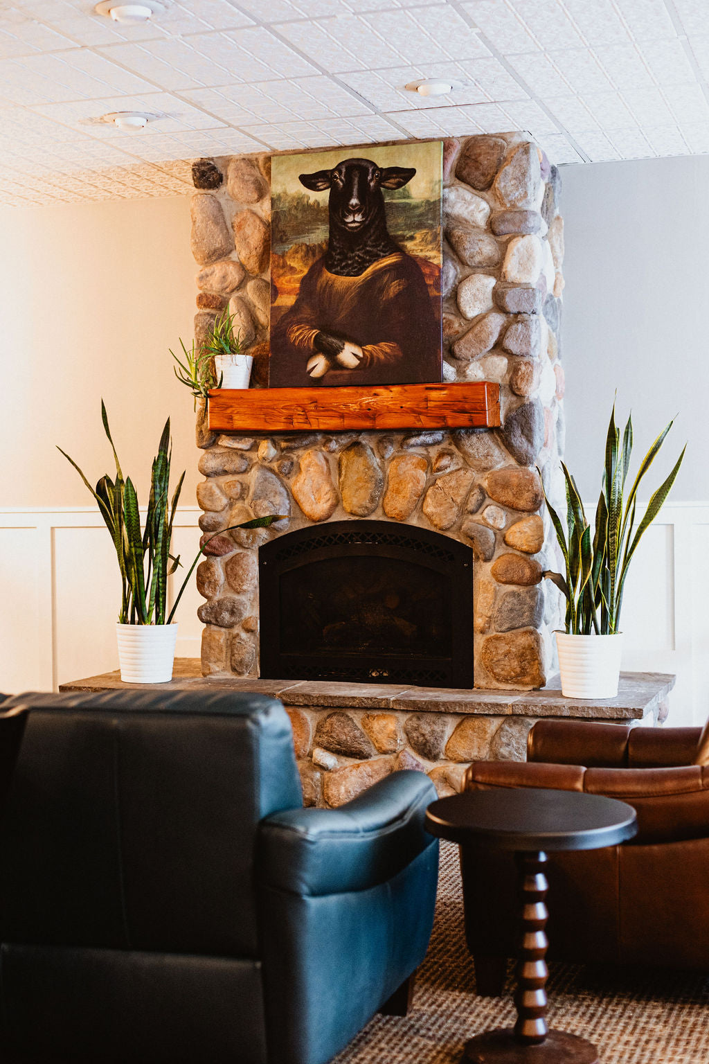 A cozy and inviting stone fireplace area with a unique "Mona Lisa" styled portrait of a black sheep hanging above the wooden mantel. Flanked by potted snake plants on either side, the scene is complemented by comfortable leather chairs and a small side table, creating a warm and relaxing atmosphere.